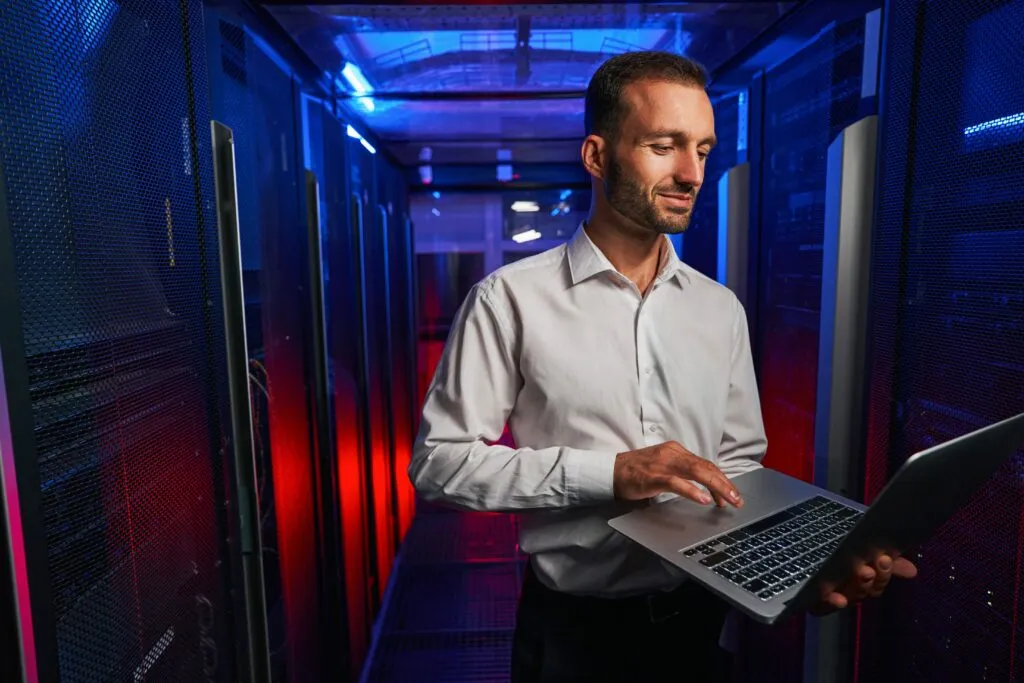 Cloud Platform: Caucasian programmer is looking to the screen of laptop while standing near the server rack