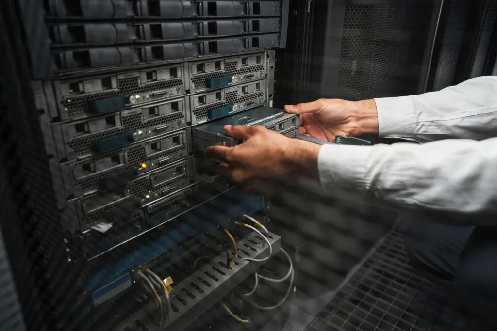 Cloud Platform: Caucasian programmer is looking to the screen of laptop while standing near the server rack